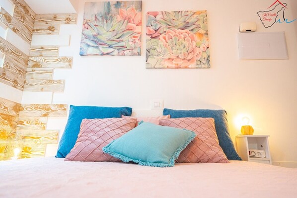 Un dormitorio con cama, almohadas, decoración en la pared y una mesita de noche con lámpara.