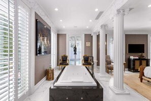 A luxurious room with a large bathtub, two chairs, a desk, and a television.