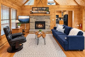 A cozy cabin interior with a stone fireplace, a blue sofa, a leather chair, and a television. - Fall Retreat | Private Hot Tub + Free Tickets (Sevierville)