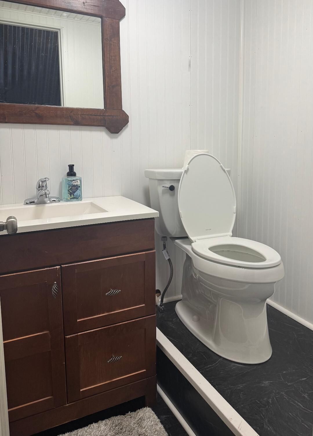 A bathroom with a white toilet, a wooden vanity with drawers, a mirror, and a window with blinds.
