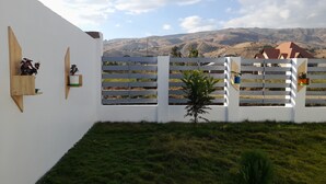Eine weiße Wand mit Holzregalen, auf denen Blumentöpfe stehen, ein weißer Zaun mit horizontalen Latten und eine Rasenfläche mit einem kleinen Baum.