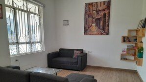 A living room with a sofa, a glass coffee table, a painting on the wall, and a wooden bookshelf.