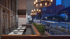 An outdoor dining area with multiple tables and chairs, a wooden bench, and a view of a city street at dusk.