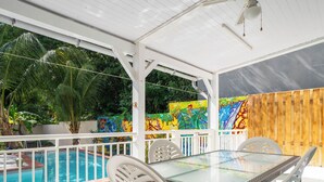 Un patio al aire libre con una mesa de vidrio y sillas blancas, una piscina y murales coloridos en las paredes.