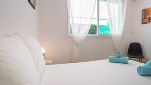 Un dormitorio con cama, ventana con cortinas translúcidas, una silla y aire acondicionado instalado en la pared.