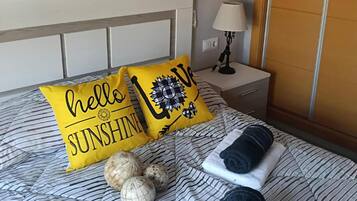 Una cama bien hecha con ropa de cama a rayas, dos almohadas con el texto 'hello sunshine' y una lámpara y un espejo en la pared.