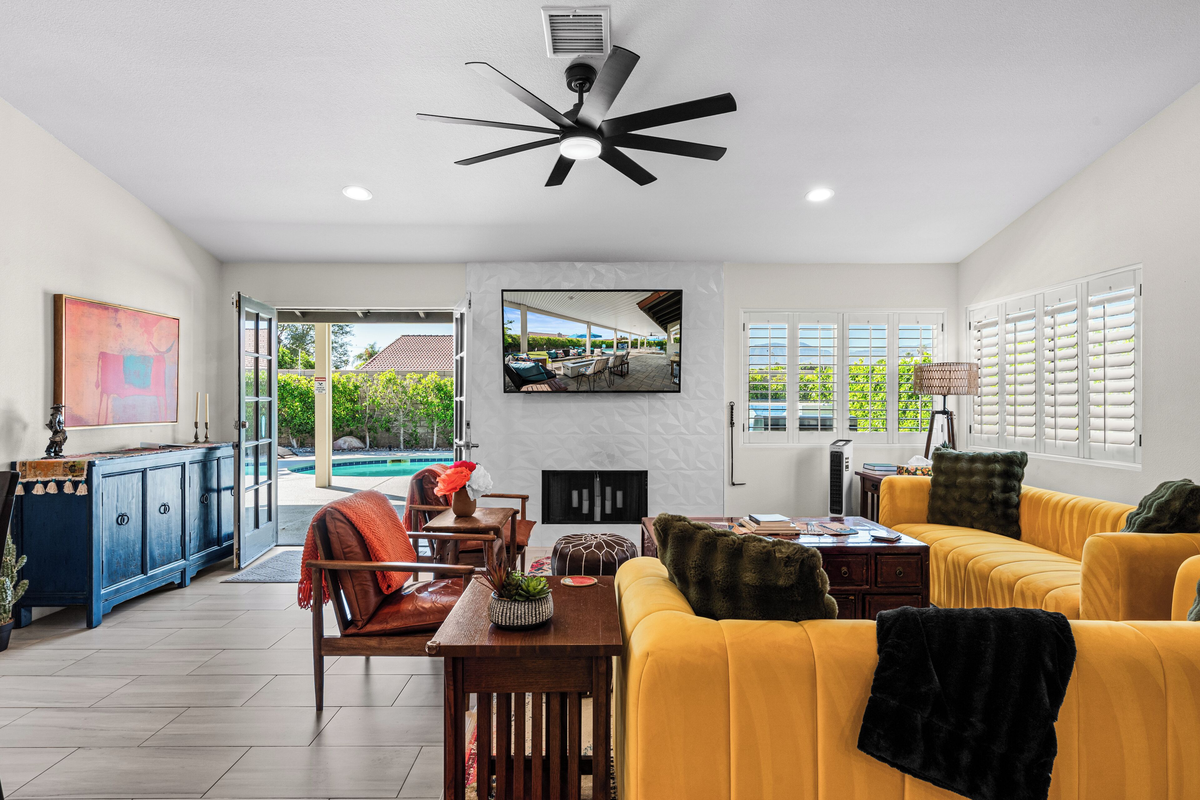 Uma sala de estar moderna com um sofá amarelo, um ventilador de teto, uma TV fixada na parede e vista para o exterior.