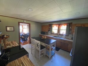 Une cuisine spacieuse avec des meubles en bois, un plan de travail noir et un réfrigérateur. On y trouve un coin repas avec une table et des chaises, ainsi qu’une fenêtre avec des rideaux.