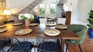 A dining area with a wooden table set for six, surrounded by chairs, a staircase, and a view of greenery outside.