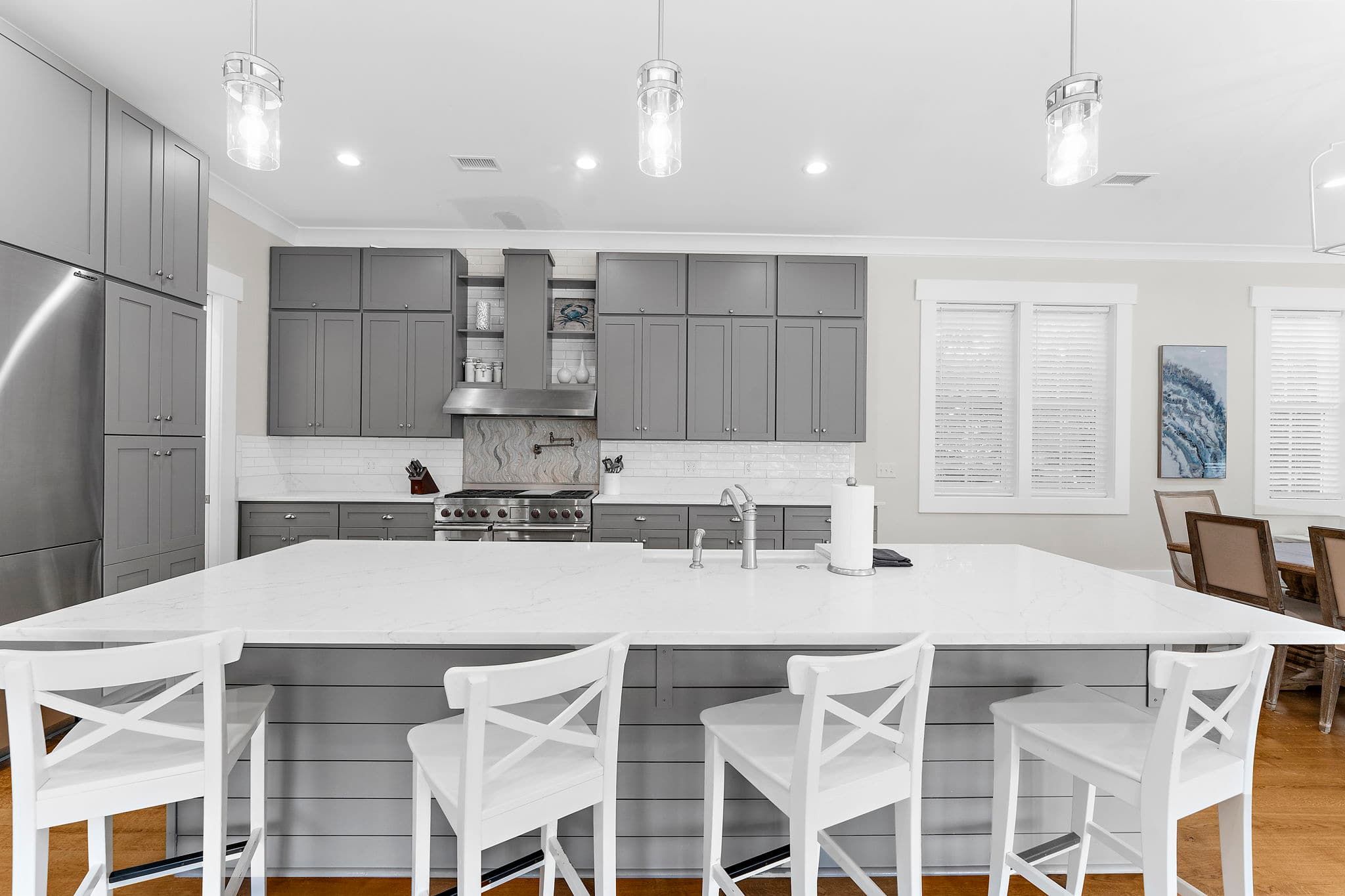 A modern kitchen with grey cabinets, a white island with bar stools, stainless steel appliances, and pendant lighting.