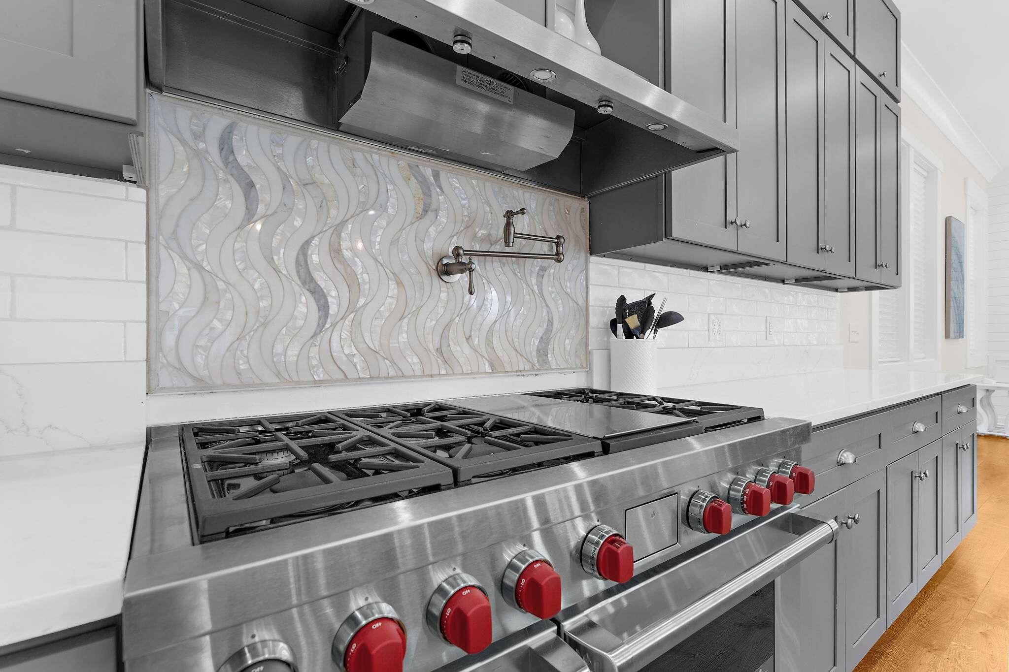 A kitchen with a stainless steel range hood, a tiled backsplash, and a gas stove with red knobs.
