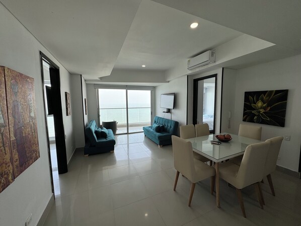 A modern living room with a glass door leading to a balcony, a dining area with a glass table, and a large painting on the wall. - Apartamentos Palmetto Eliptic ICDI (Cartagena)