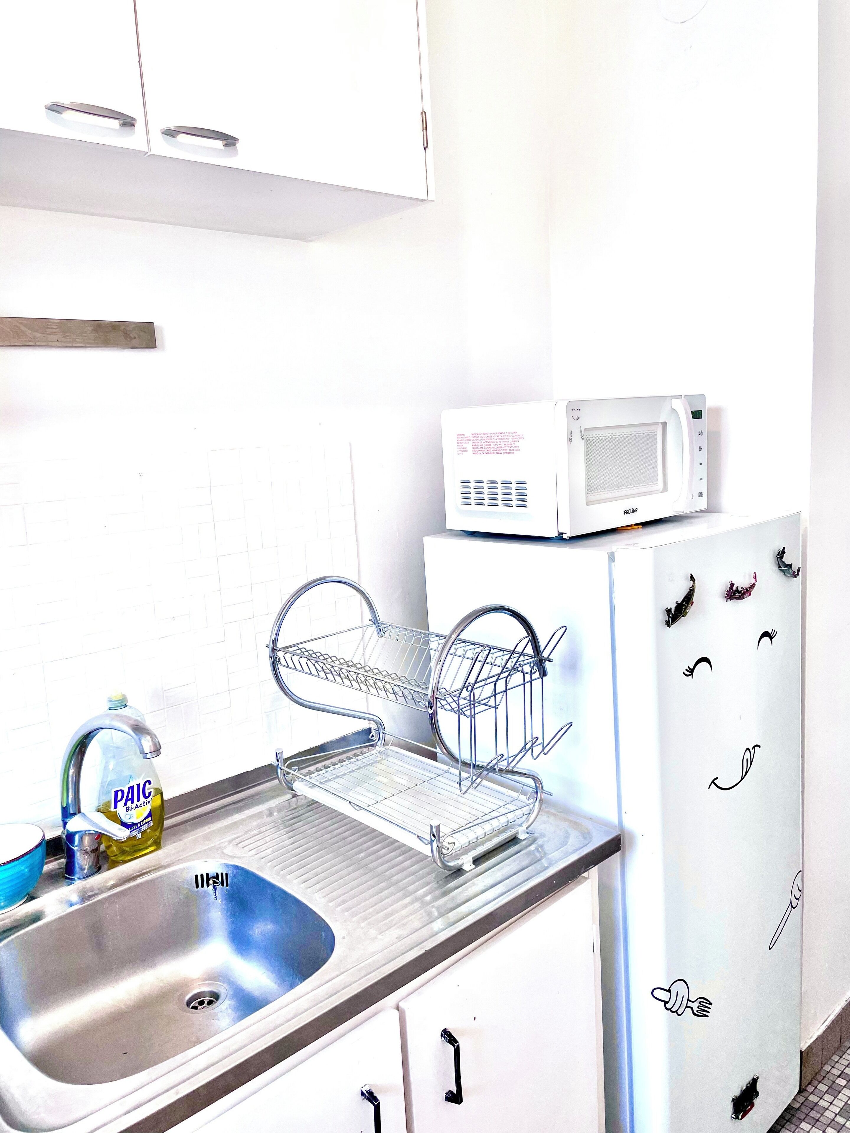 A kitchen with a stainless steel sink, a dish drying rack, a microwave, and a refrigerator with a face decal.