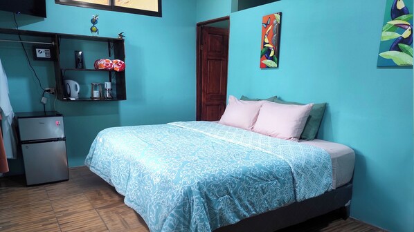 A bedroom with a bed, a small kitchenette, a wooden door, and a wall with a painting. - Hotel vista Bonita- Tucan Room (Parrita)
