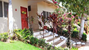 Une maison avec une porte rouge, un escalier menant à l’entrée et un jardin où poussent différentes plantes et fleurs.