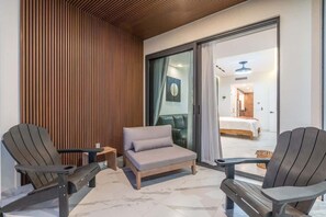 A modern interior with a sofa and two Adirondack chairs, a wooden slatted wall, and a glimpse of a bedroom through a glass door.