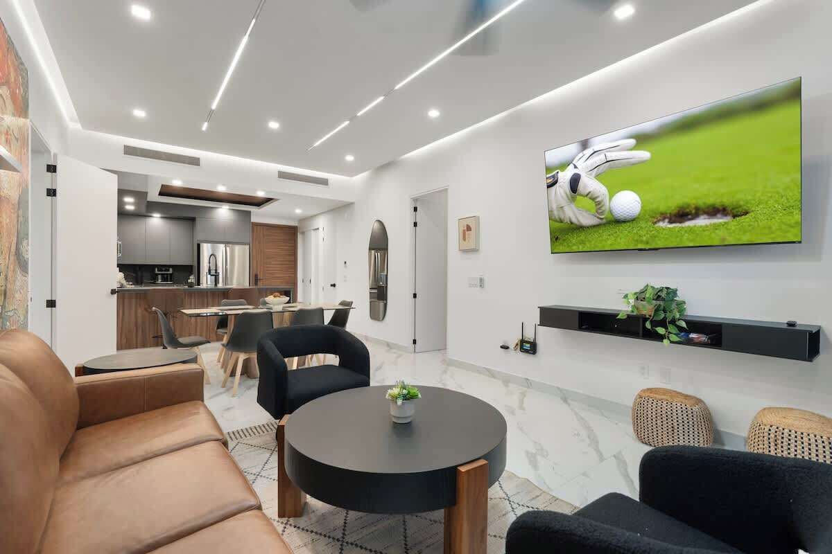 Una sala moderna con televisor de pantalla plana, sofás de cuero, una mesa de centro redonda y una cocina al fondo.