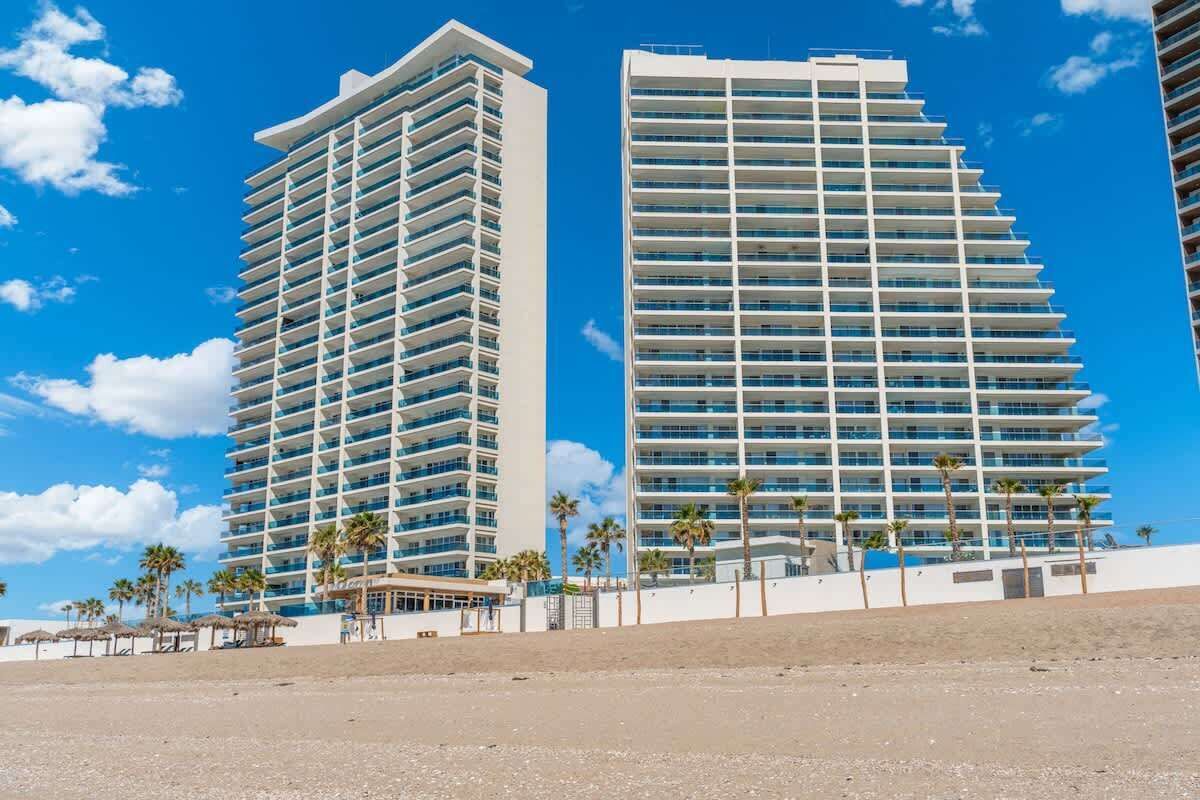 Two high-rise buildings with multiple balconies, a sandy beach in the foreground, and palm trees.