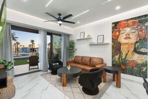 A modern living room with a sofa, two armchairs, and a coffee table. There is a large window with a view of palm trees and a swimming pool.