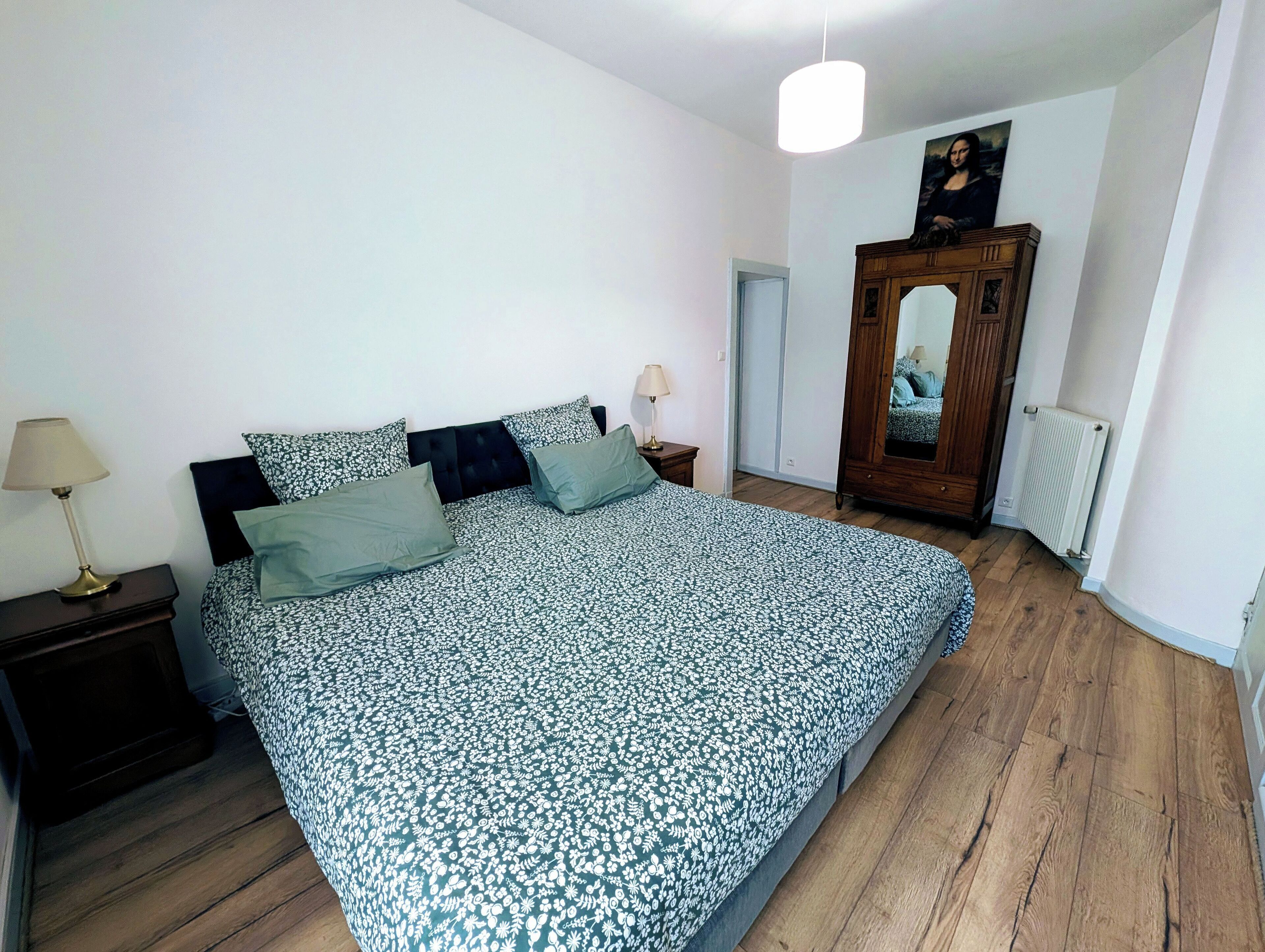 Un dormitorio con una cama grande, un armario de madera, una mesita de noche con una lámpara y un espejo sobre el armario.