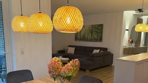 Een moderne eetruimte met een houten tafel, zwarte stoelen en een bloevas. Drie hanglampen met een geweven ontwerp. Op de achtergrond is een grijze bank en een schilderij te zien.