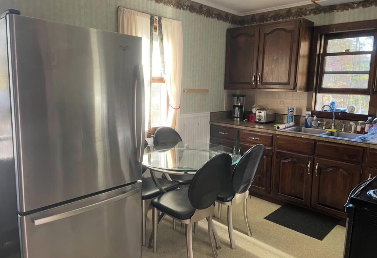 A compact kitchen with a stainless steel refrigerator, a glass dining table with four chairs, wooden cabinets, and a window with curtains.