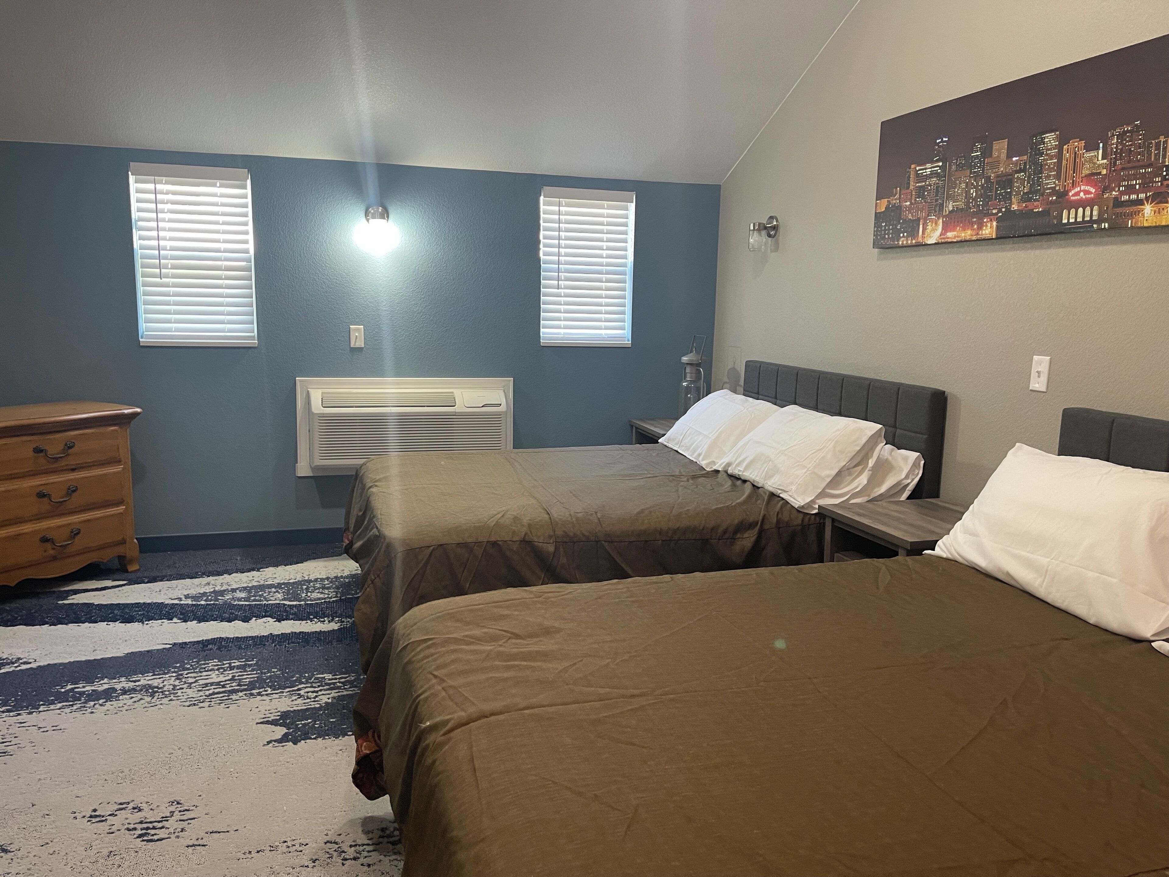 A bedroom with two beds, a dresser, a heater, and two windows with blinds.