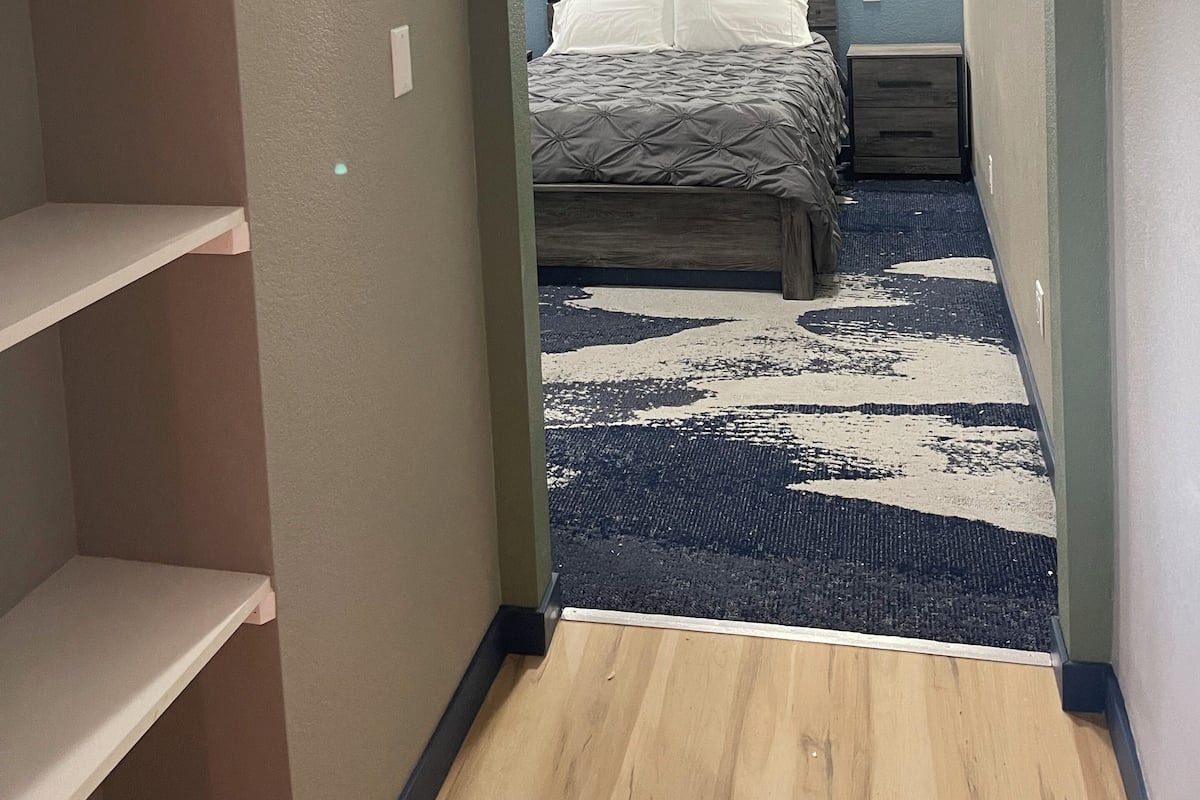 A narrow hallway leading to a bedroom with a bed, a nightstand, and a wall-mounted light.