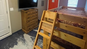 Quarto com beliche, televisão, cómoda de madeira e porta com moldura vermelha.