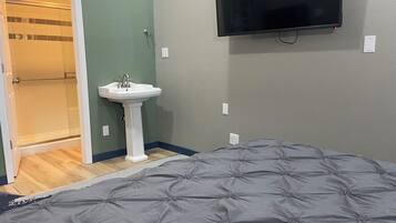 A bedroom with a bed, a wall-mounted TV, a pedestal sink, and a shower.