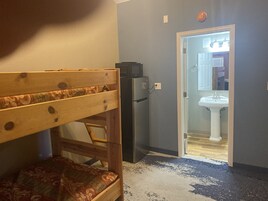 Una habitación con literas, un refrigerador y un baño visible a través de una puerta abierta.