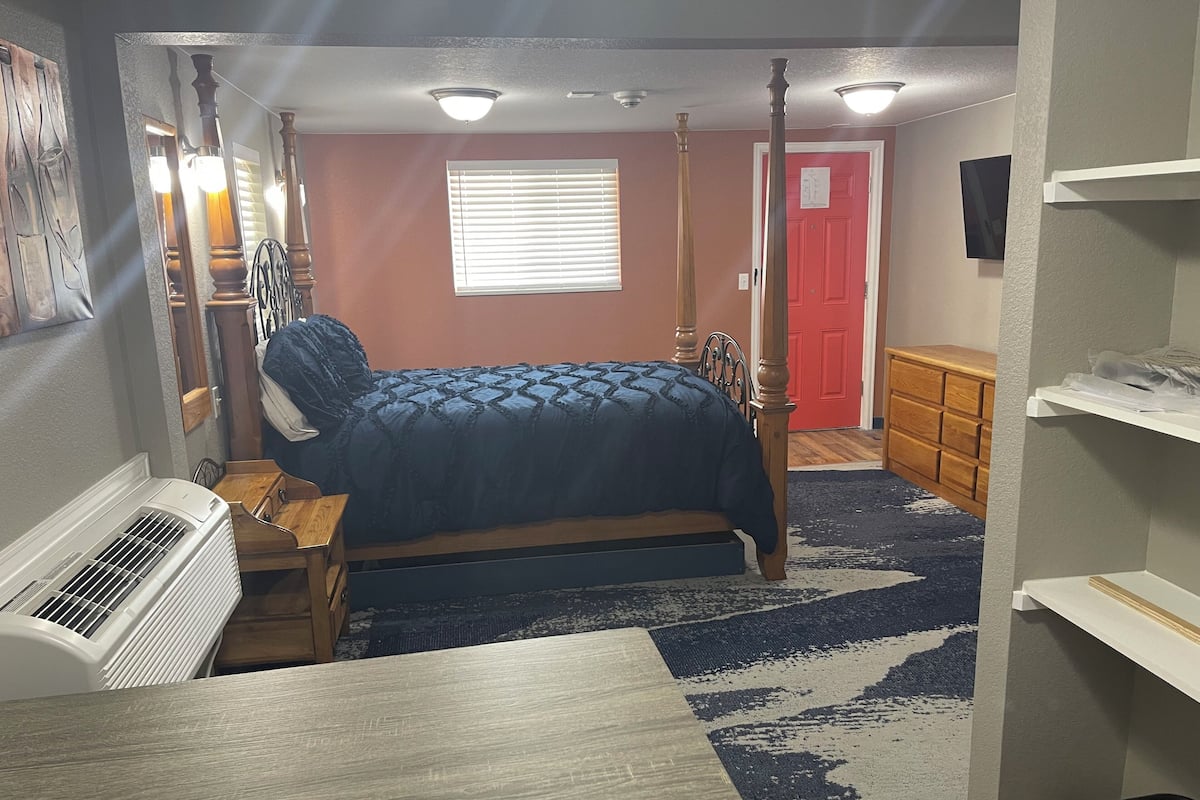 A bedroom with a bed, a heater, a dresser, a red door, and a window with blinds.
