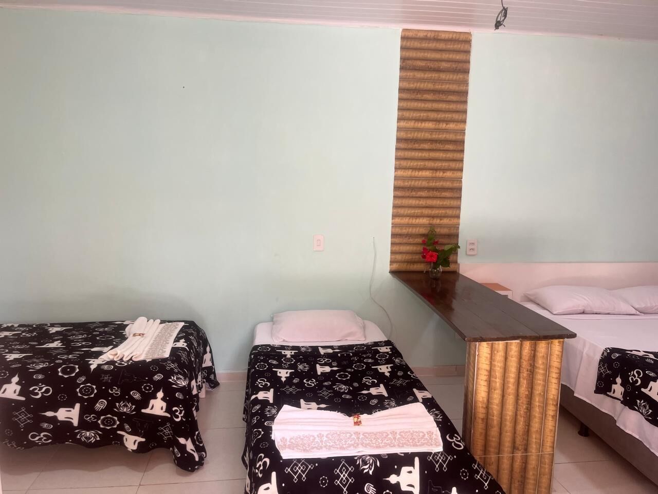 A room with three beds, each with a black and white patterned bedspread. There is a wooden bench in the center, and a small table with a plant on it.