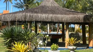 A thatched-roof gazebo with a table and chairs, surrounded by tropical plants and palm trees. - Pousada Oasis Beach Club (Canavieiras)