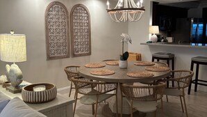 A dining area with a round table, wicker chairs, a chandelier, and a kitchen in the background. - Spacious Top-Floor Condo w/beach access & Intracoastal Views (Indian Shores)