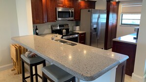 A kitchen with granite countertops, stainless steel appliances, wooden cabinets, and a bar stool. - Spacious Top-Floor Condo w/beach access & Intracoastal Views (Indian Shores)