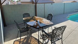 Eine überdachte Terrasse mit einem Glastisch und Metallstühlen, im Hintergrund ein Pool und eine Backsteinmauer.