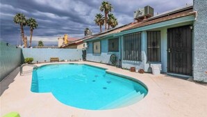 Ein Pool mit geschwungener Kante, umgeben von einer hellen Terrasse. Im Hintergrund sind Palmen und ein Gebäude mit blauer Fassade und schwarzem Eingang zu sehen.
