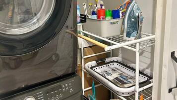 A laundry area with a front-loading washing machine, a drying rack, and shelves stocked with cleaning supplies.