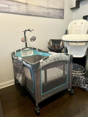 Un parc pour bébé avec des roulettes, un siège pour bébé et une petite cuvette blanche dans une pièce dont le sol est en bois et où l’on peut voir un tableau accroché au mur.