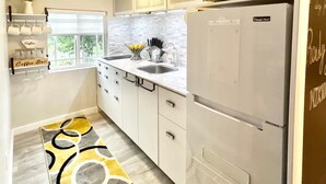 Una cocina moderna con armarios blancos, microondas, refrigerador, barra de café y ventana con vista a zonas verdes.