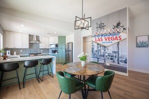 A modern kitchen with white cabinets, a central island with bar stools, and a dining area with a round table and green chairs. - Stylish 4BR Oasis w/ Pool, Spa, Firepit, Ping Pong (North Las Vegas)