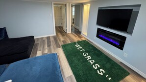 Ein modernes Wohnzimmer mit einem an der Wand befestigten Flachbildfernseher, einem blauen Sofa und einem grünen Teppich mit dem Aufdruck 'WET GRASS'.