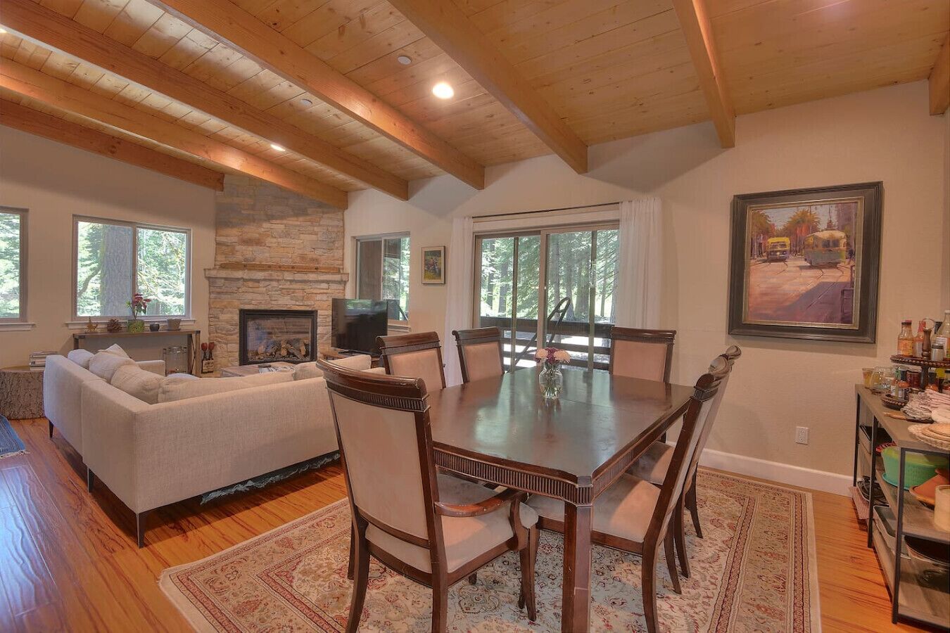 A spacious living room with a fireplace, a dining area with a wooden table and chairs, and large windows offering natural light and views of the outdoors.