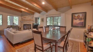 A spacious living room with a fireplace, a dining area with a wooden table and chairs, and large windows offering natural light and views of the outdoors. - Palisades-Alpine Home w/ Hot Tub (Alpine Meadows)