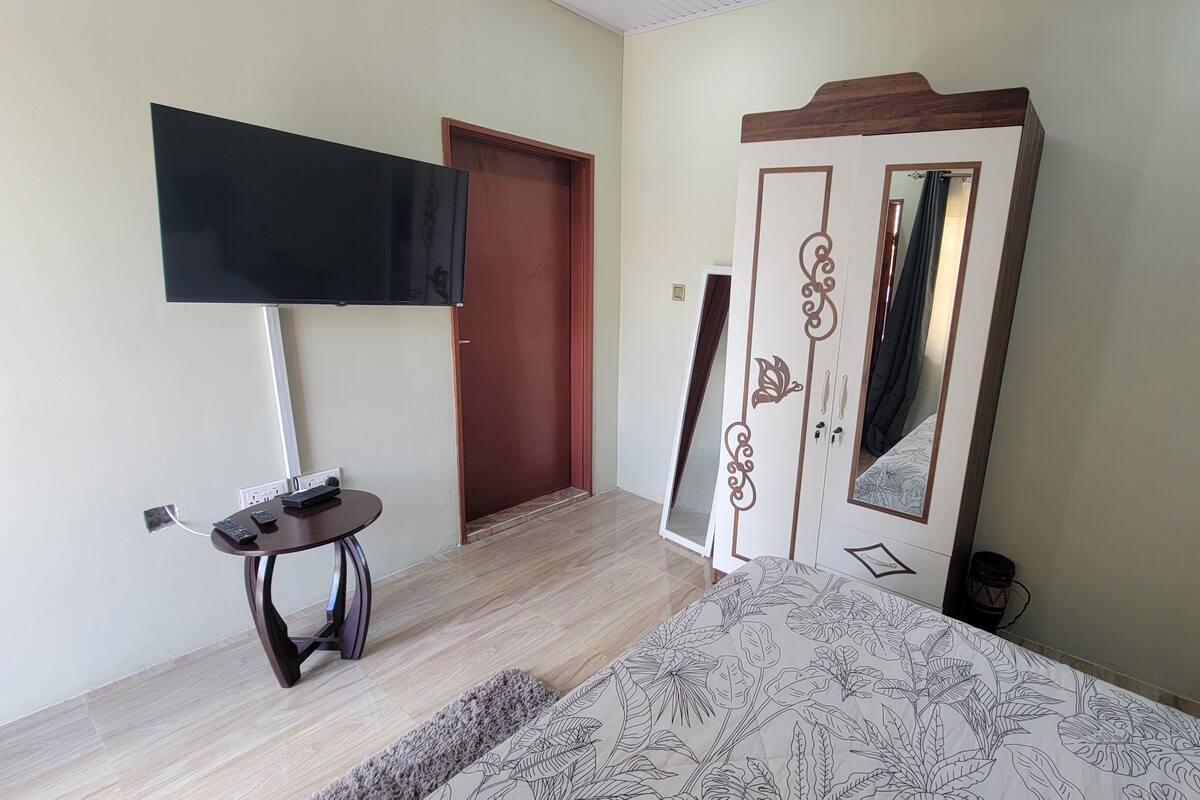 A hotel room with a flat-screen TV mounted on the wall, a wooden door with decorative panels, a bed with a patterned bedspread, and a small round table with a remote control.