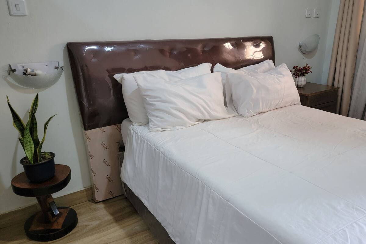 A neatly made bed with white bedding, a dark brown headboard, and a small bedside table with a vase of flowers.