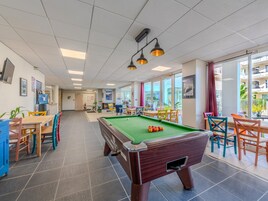 Una mesa de billar en una habitación luminosa con amplios ventanales, un área de comedor con sillas de colores, y una cocina visible al fondo.