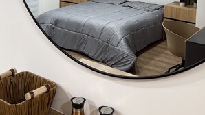 Un dormitorio moderno con un espejo grande, una cama con ropa de cama gris, mesitas de noche de madera y una pequeña silla de madera.