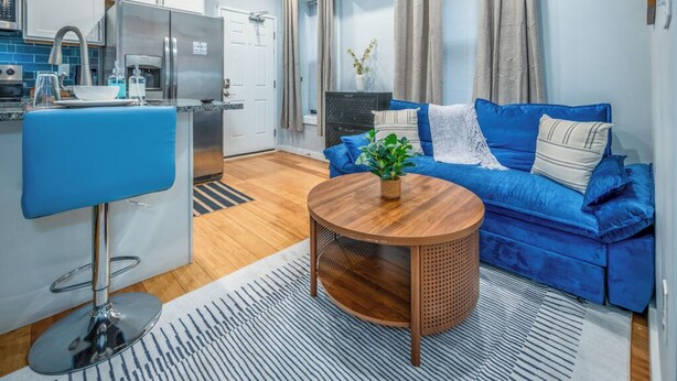 A compact living space with a kitchenette, a blue sofa, a round wooden coffee table, and a striped rug. - OTR by TQL+Smart TVs+King Bed+Wifi+Sleeps 3+1st Fl (Cincinnati)